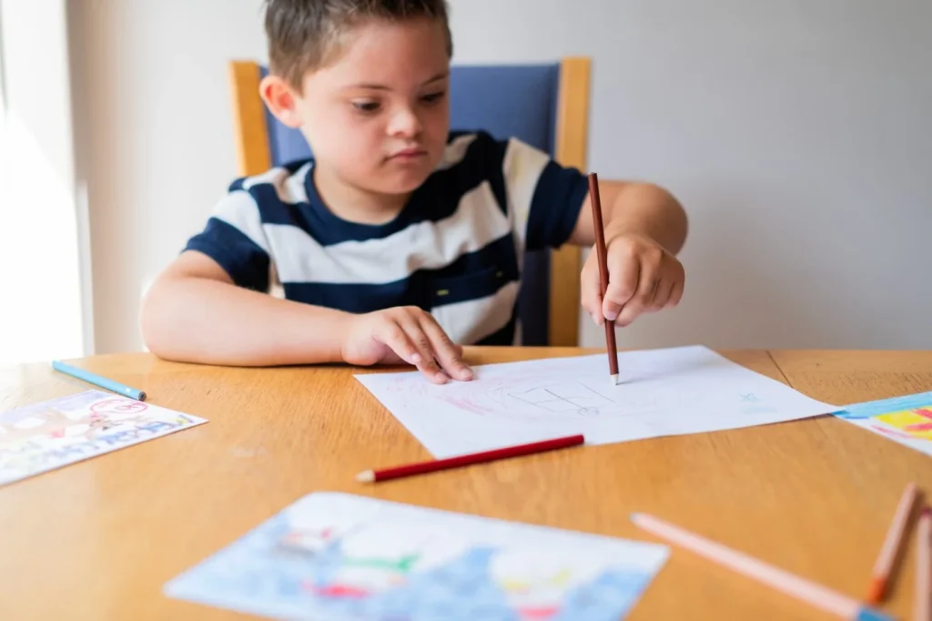 Young disabled child with down syndrome drawing on paper with pencils